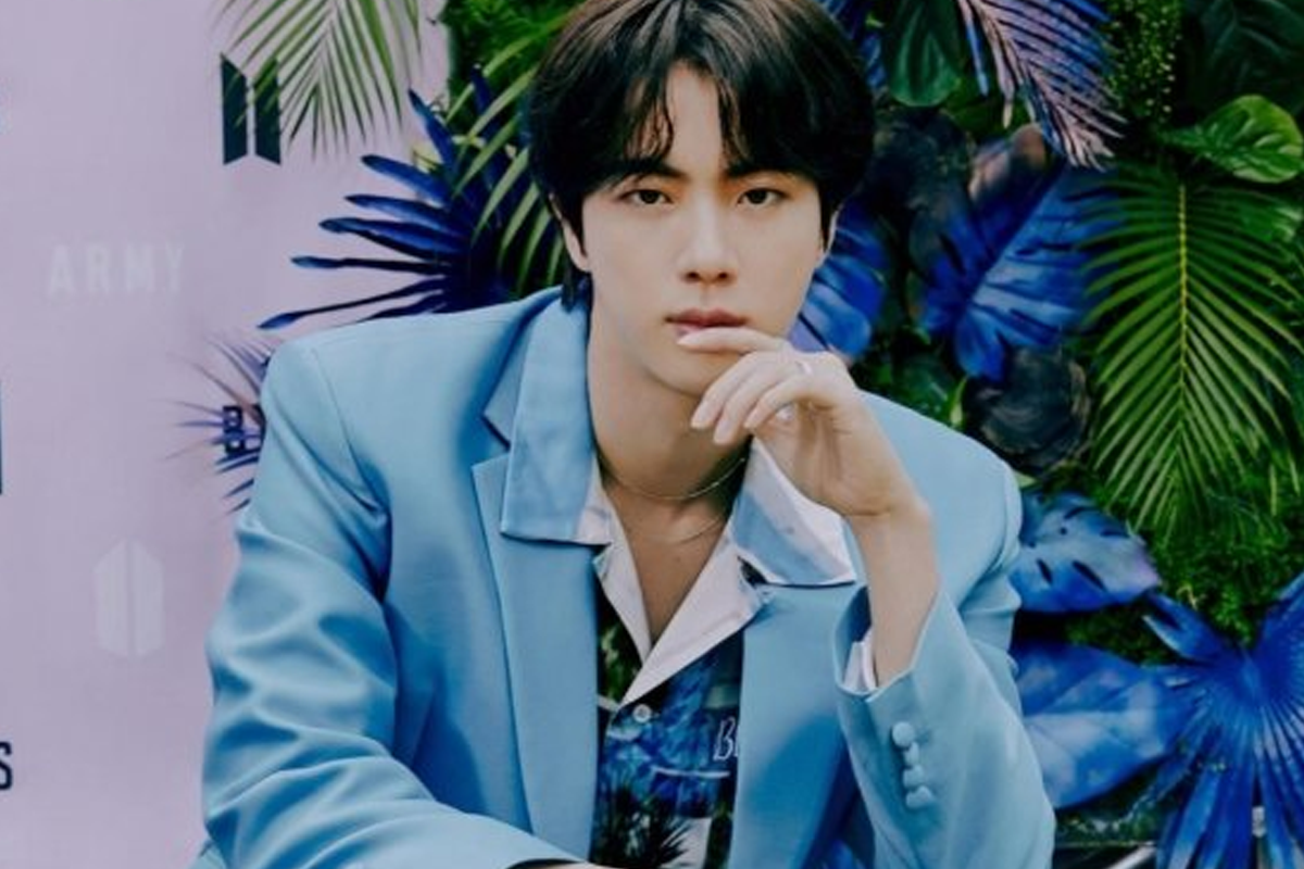 BTS成員JIN身穿藍色服裝靠在光亮背景前，展現出清新的外觀。
