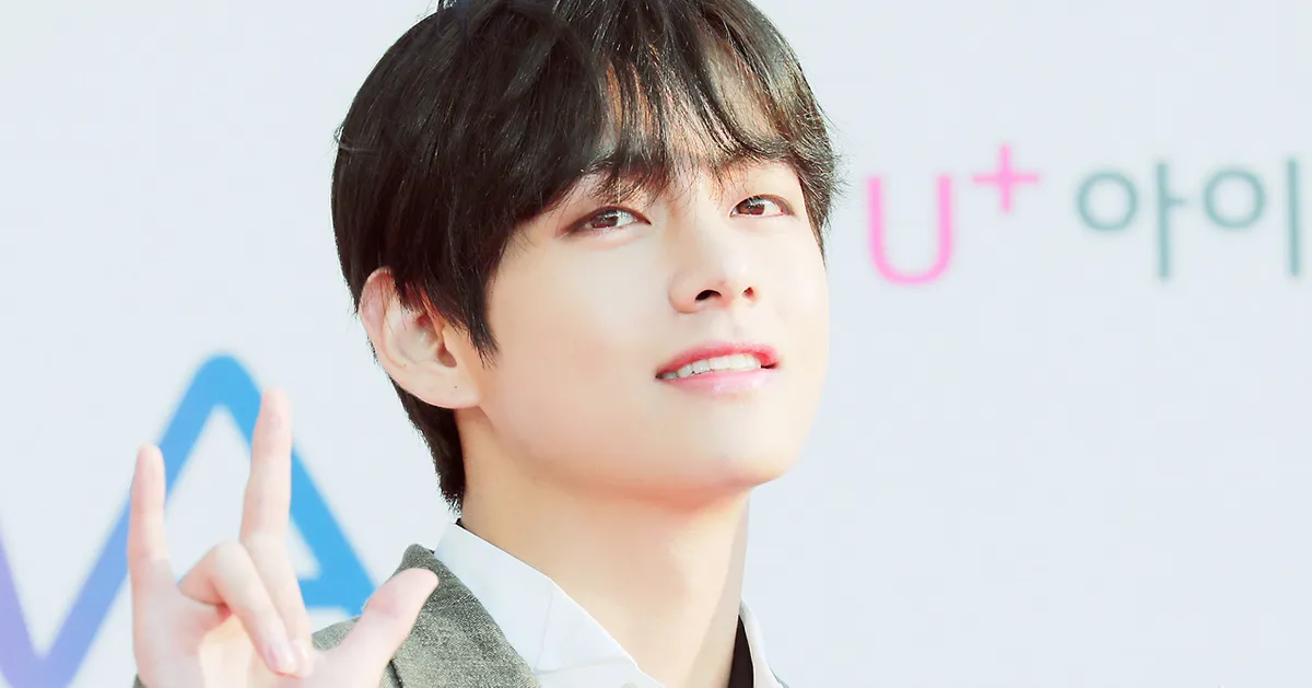 BTS成員之一以獨特的淡藍色頭髮坐在室內，穿著粉紅色襯衫，場景充滿生活的舒適感。