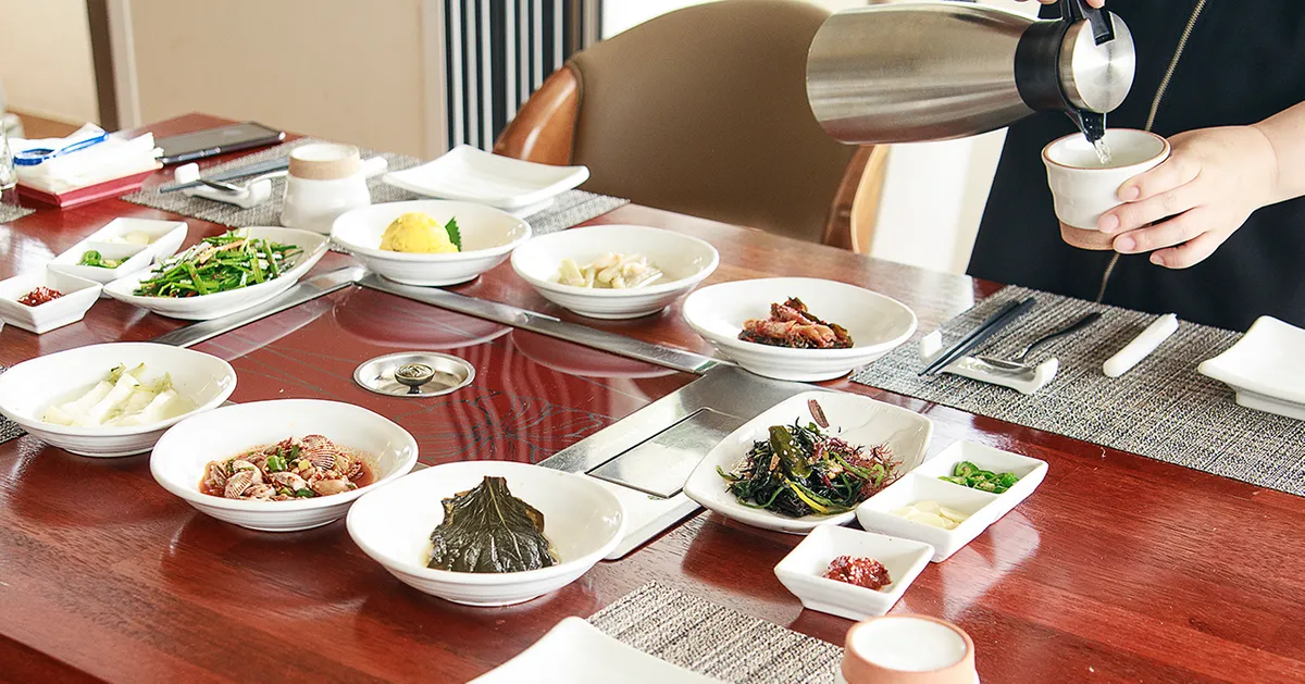 韓國員工將傳統餐桌上的飲用水注入杯中，背景是豐盛的韓國料理擺設