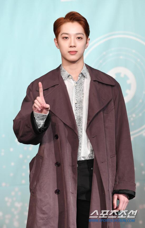 ベージュのトレンチコートを着た男性が、指で数字1を示している。スポーツ朝鮮のロゴが写真の右下にあります。背景には円の模様が描かれた淡いブルーの幕があります。