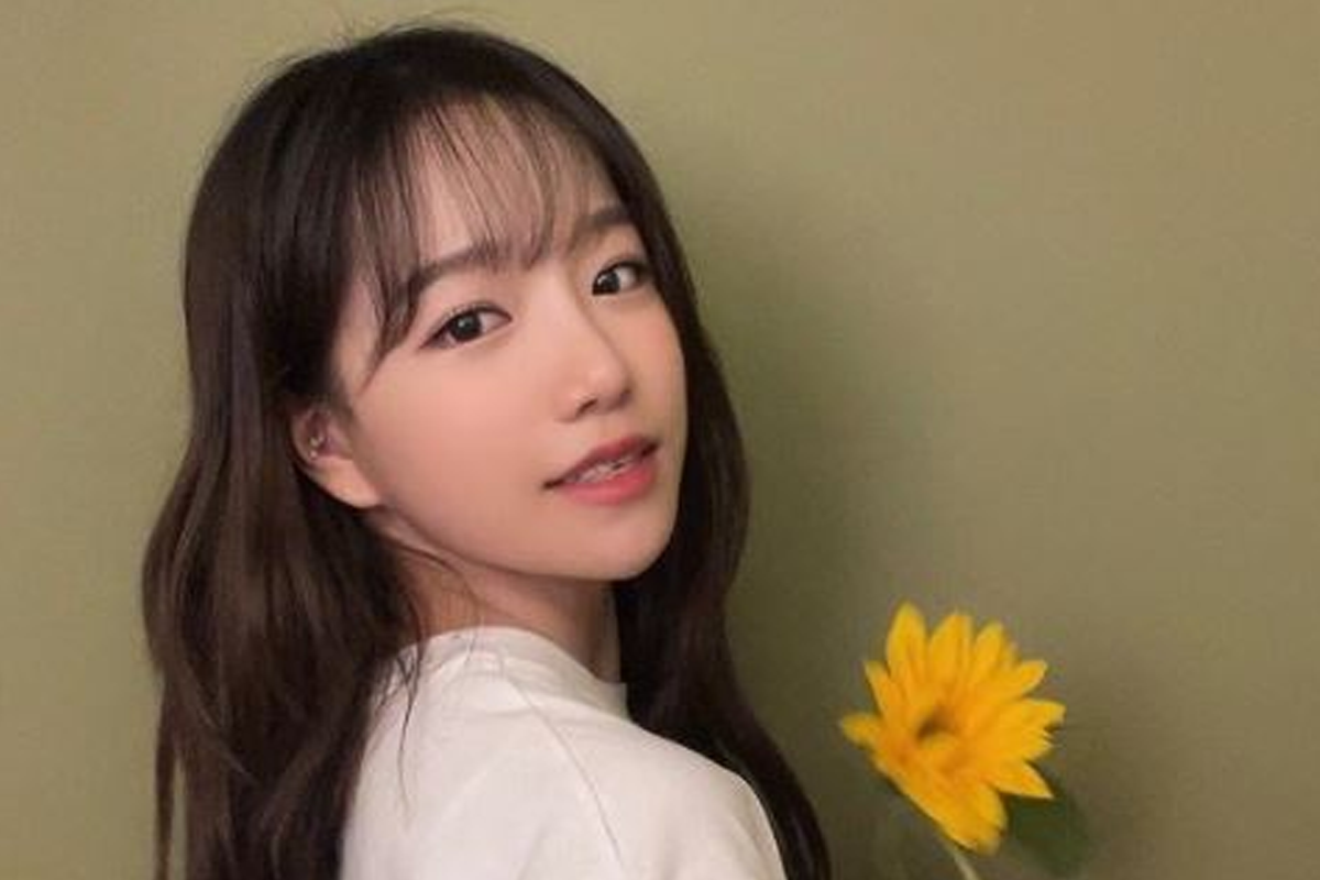 未知韓國明星身穿白色上衣攜著向日葵花,站在綠色牆前,展現自然閒適的風格。