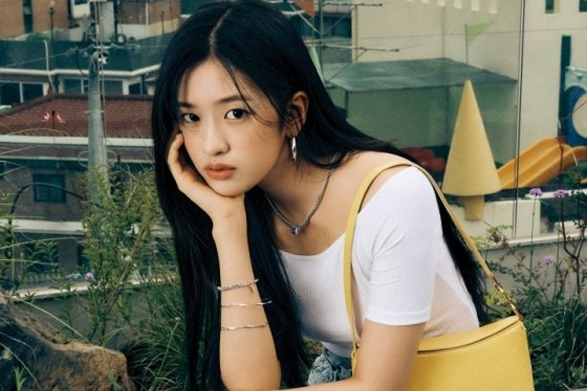 IVE成員兪真身著白色上衣,肩背黃色包包,坐在户外,背景為都市風貌,展現休閒的魅力。