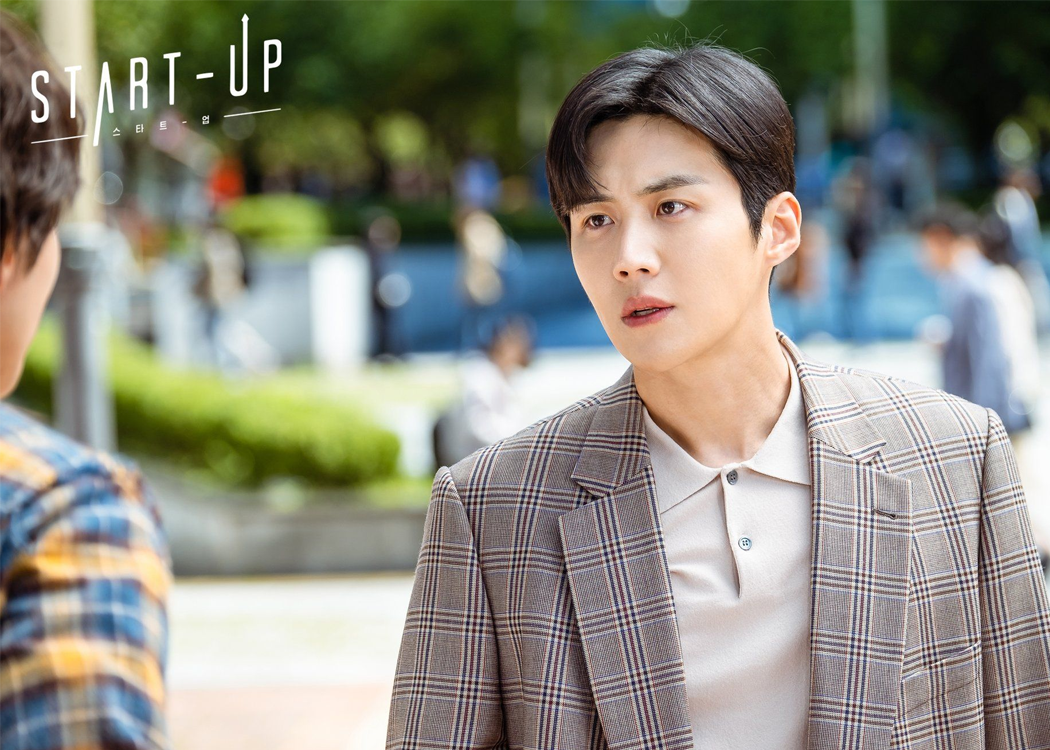 劇中角色穿著格紋西裝，置身於城市街道的場景，背景是模糊的行人和樹木。