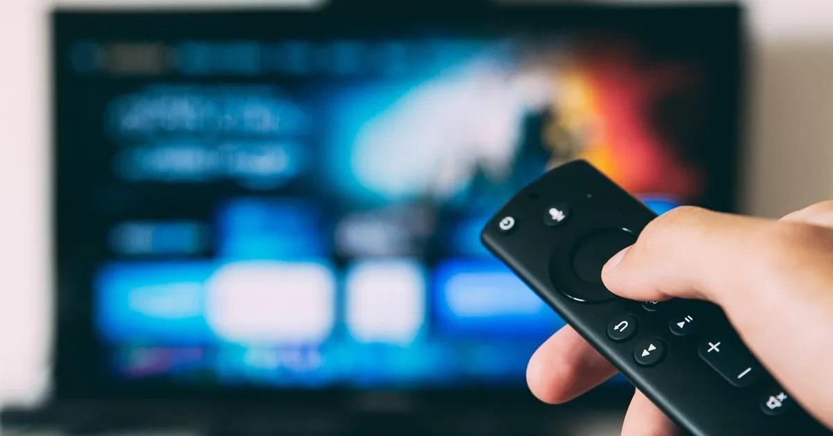 มือของผู้ถือรีโมตคอนโทรลที่กำลังชี้ไปที่หน้าจอทีวีที่แสดงเนื้อหาหลากหลาย แสดงถึงการสตรีมหรือการรับชมรายการทีวีอย่างสะดวกในบ้าน