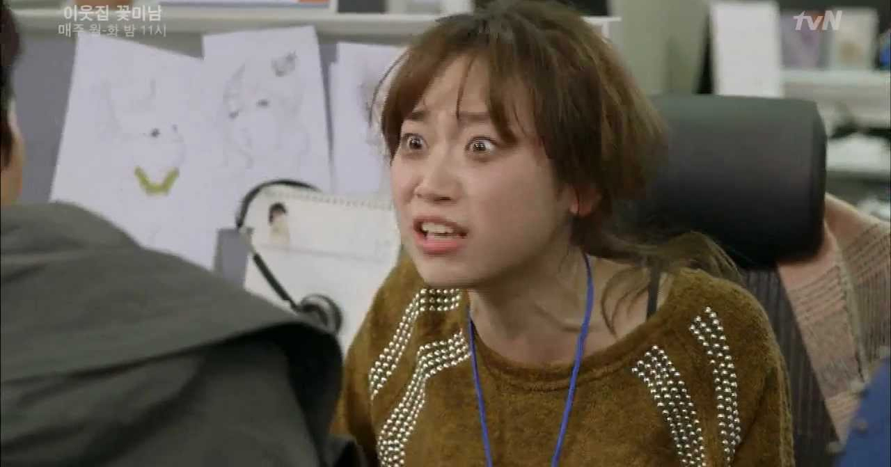 一位穿著橙色衣服和藍色識別證的韓國人在辦公室裡工作，桌上擺放著一些文件和設計畫