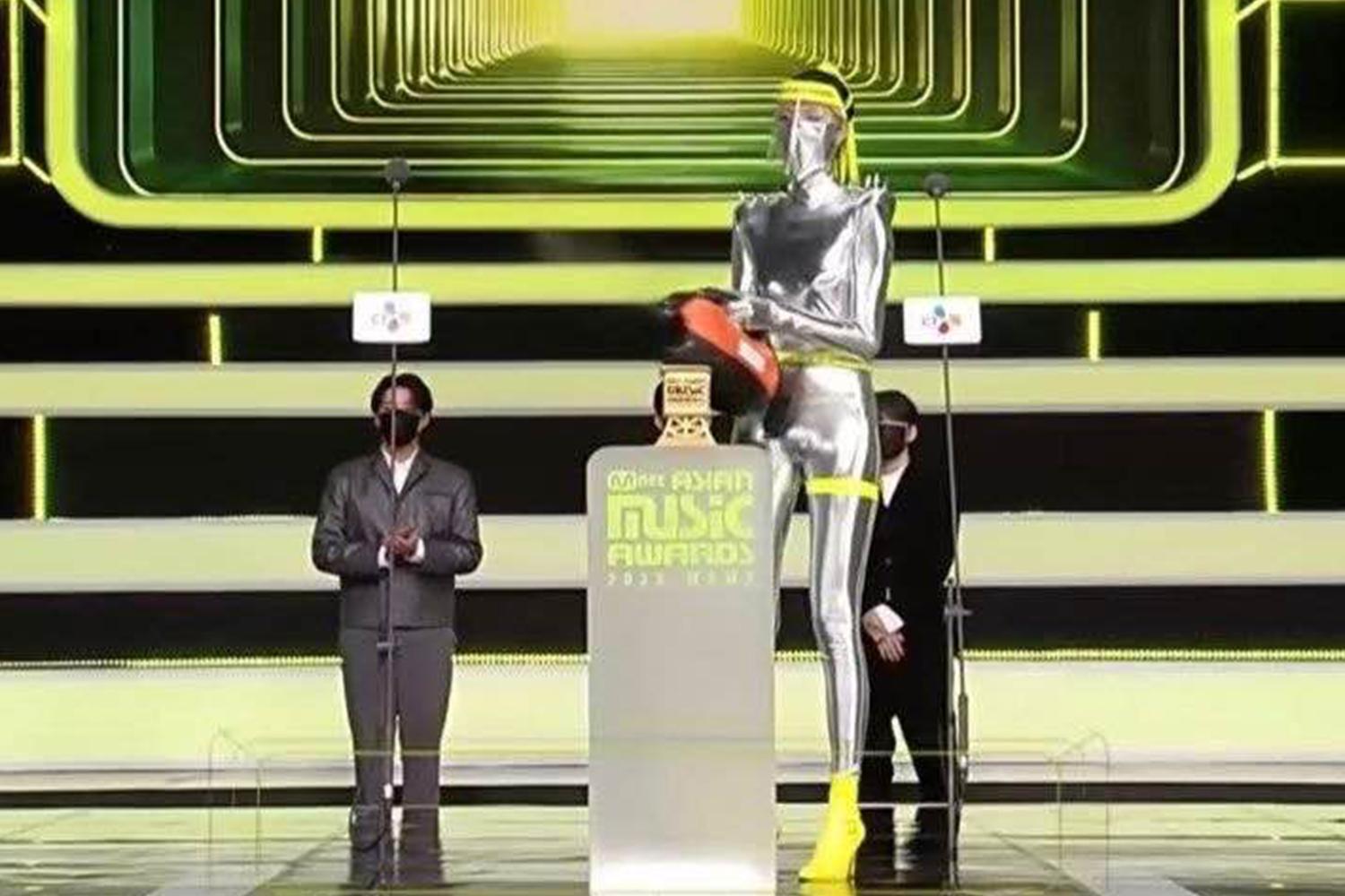 2020 MAMA 頒獎台上，一位身穿銀色華麗服裝，手持消毒噴霧的工作人員正在進行設備消毒，展現防疫措施的嚴謹性。