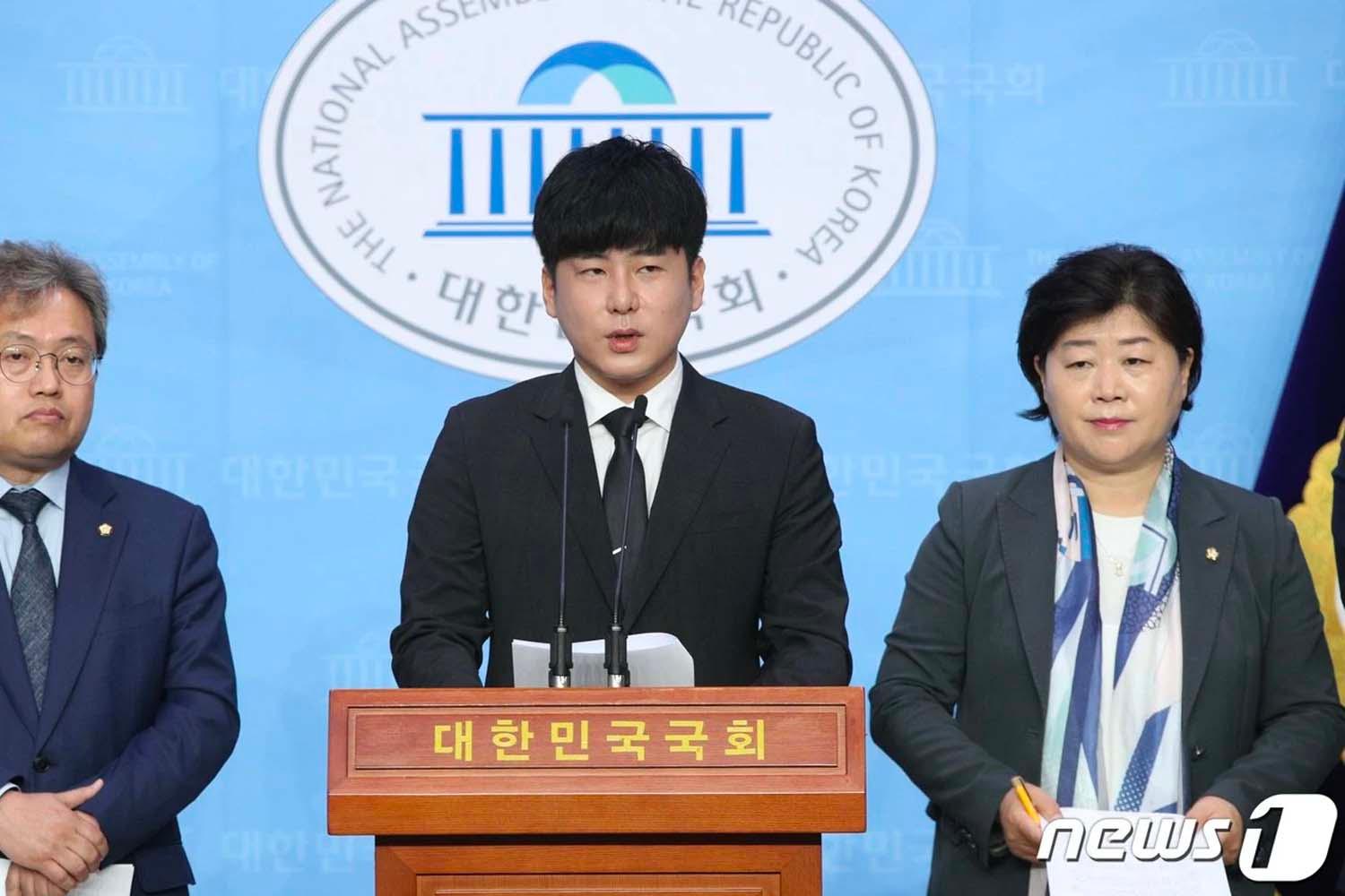 กลุ่มบุคคลยืนอยู่ในสภาพหน้าเส้นแถลงข่าวด้านหลังมีธงชาติและตราสัญลักษณ์ของสภาเกาหลีใต้