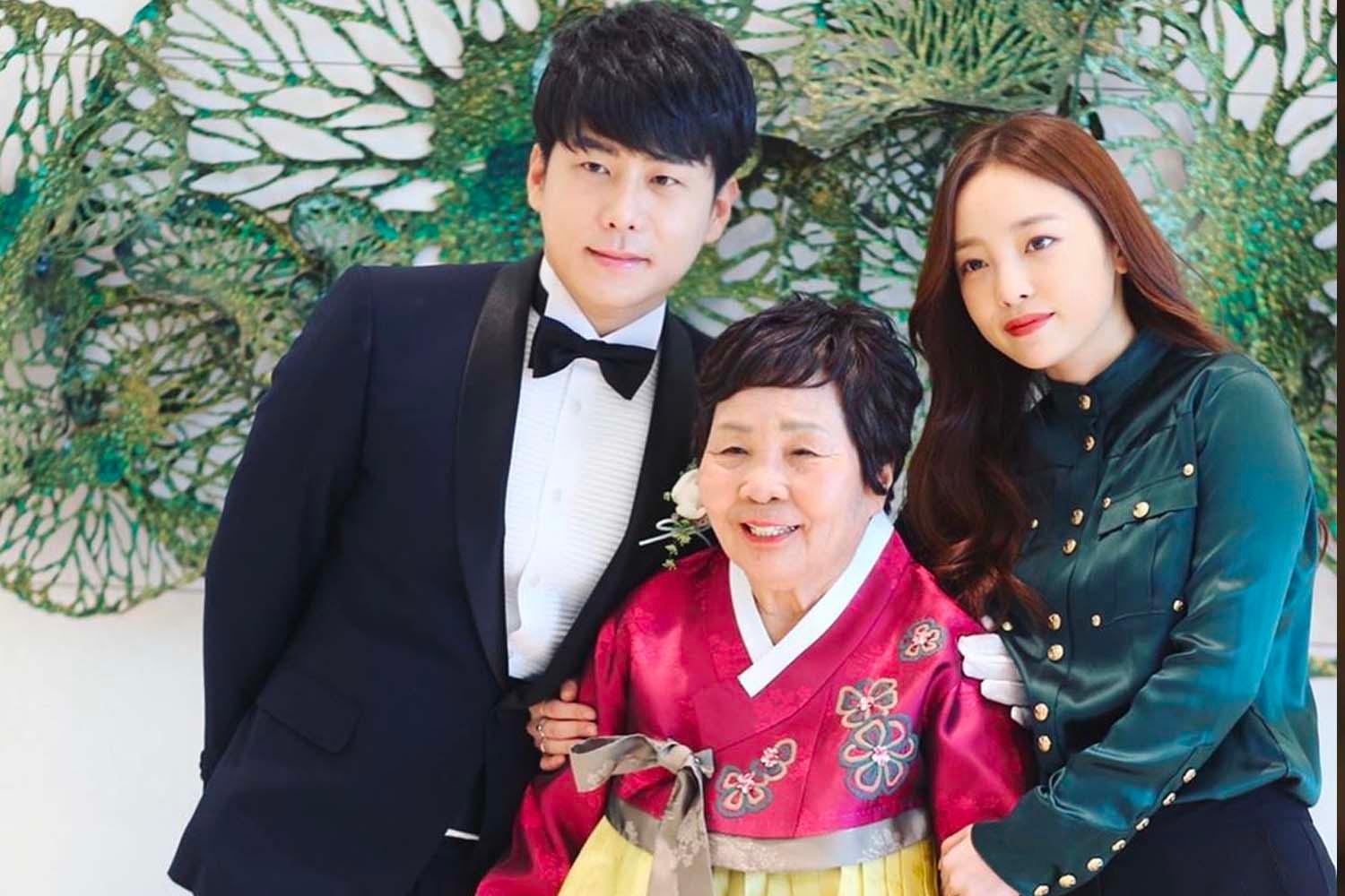 บุคคลในชุดทักซิโด้ยืนข้างผู้หญิงในชุดฮันบกสีสันสดใสและผู้หญิงในชุดสีเข้มในงานเลี้ยงแต่งงาน