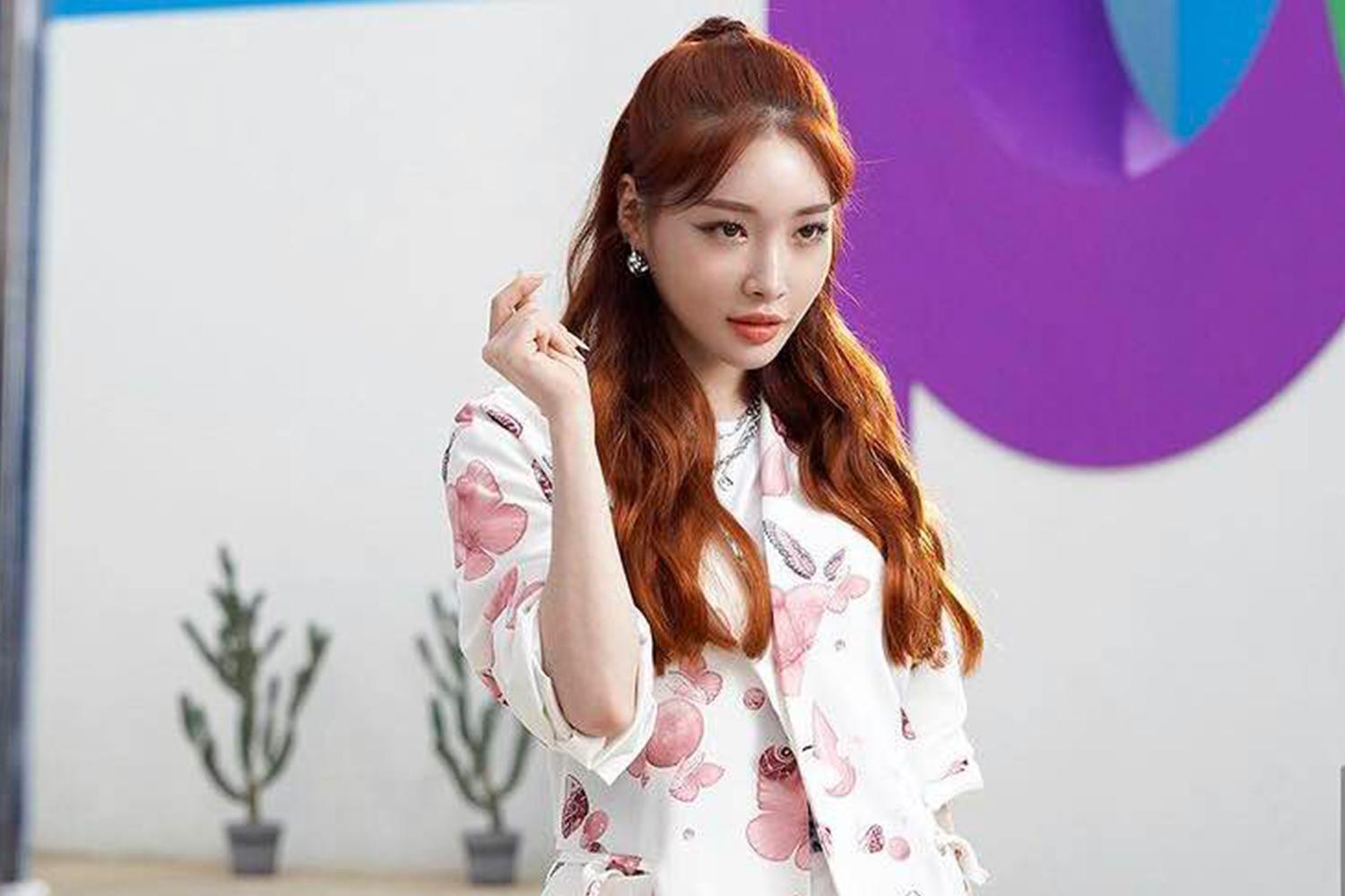 穿著白底粉紅蝴蝶花紋服飾的女歌手手勢比心，背後是室內場景