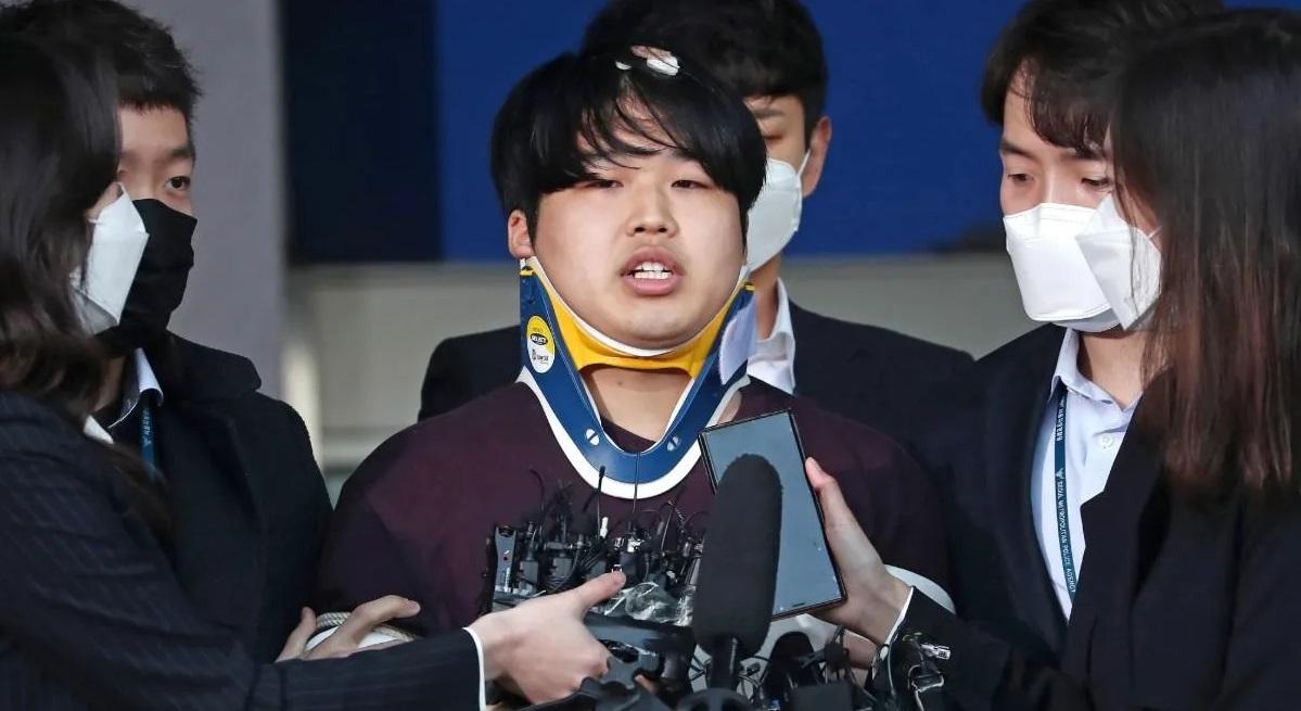 ผู้ต้องหาคดีห้อง Nth สวมเสื้อแขนยาว ใส่เฝือกคอ ยืนอยู่ระหว่างเจ้าหน้าที่ ใบหน้าถูกเบลอในกรณีที่เกี่ยวข้องกับการถูกจำคุก