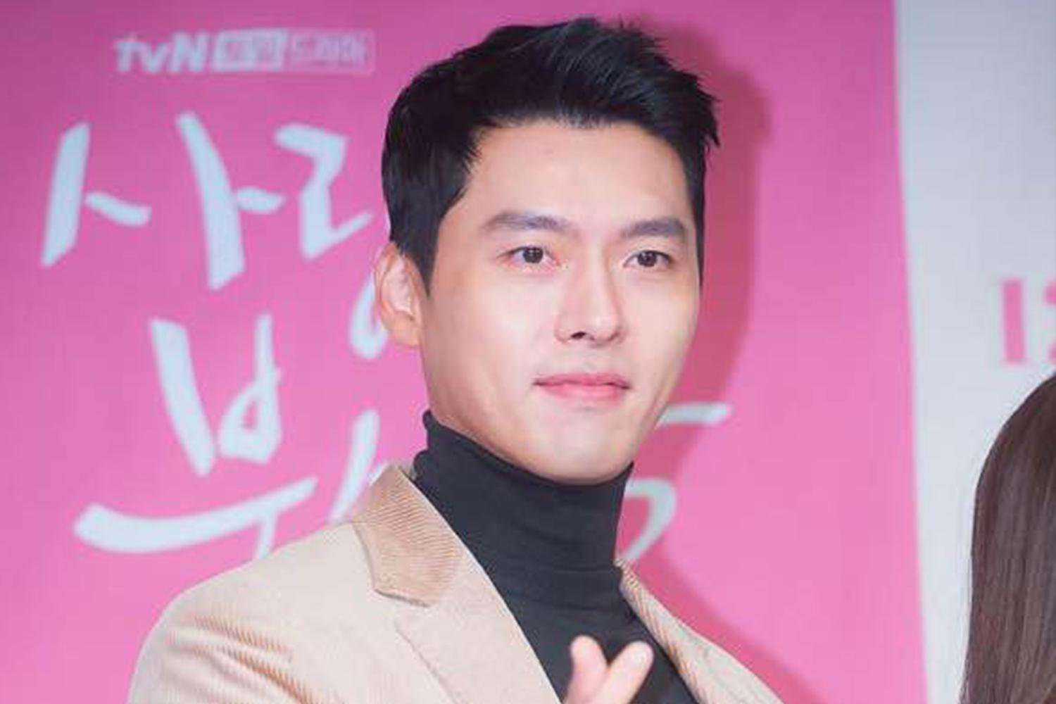 韓國男明星玄彬身穿駝色西裝外套，站在粉紅色背景前，右手做出V字手勢。