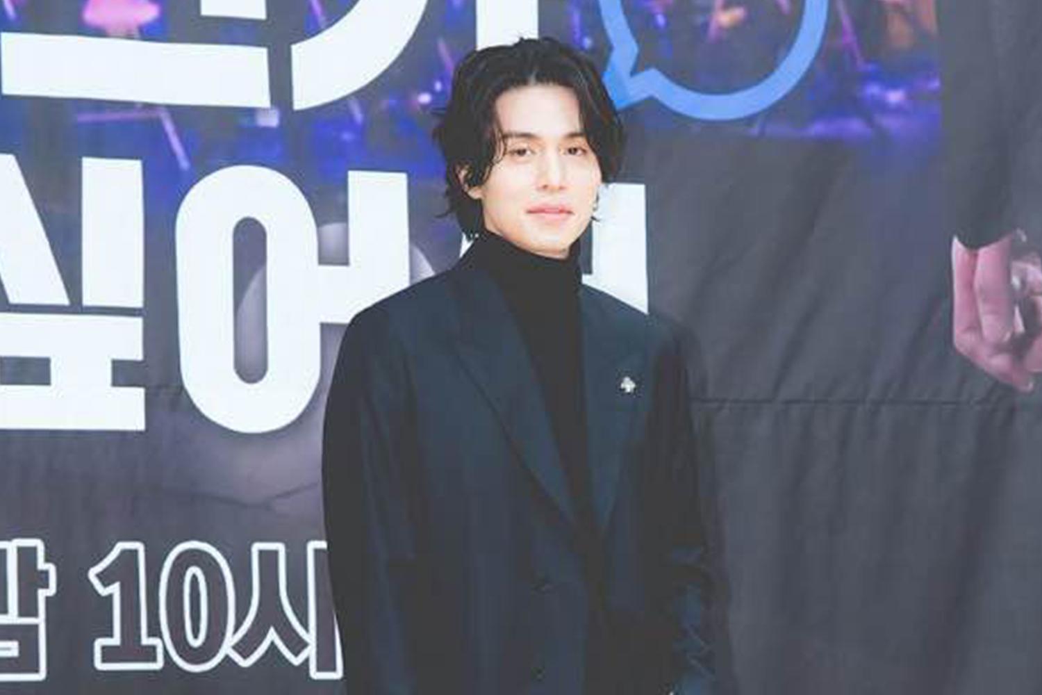 韓國男星李棟旭身穿深色西裝在活動上擺好姿勢，背景上有韓文字樣的布幕。