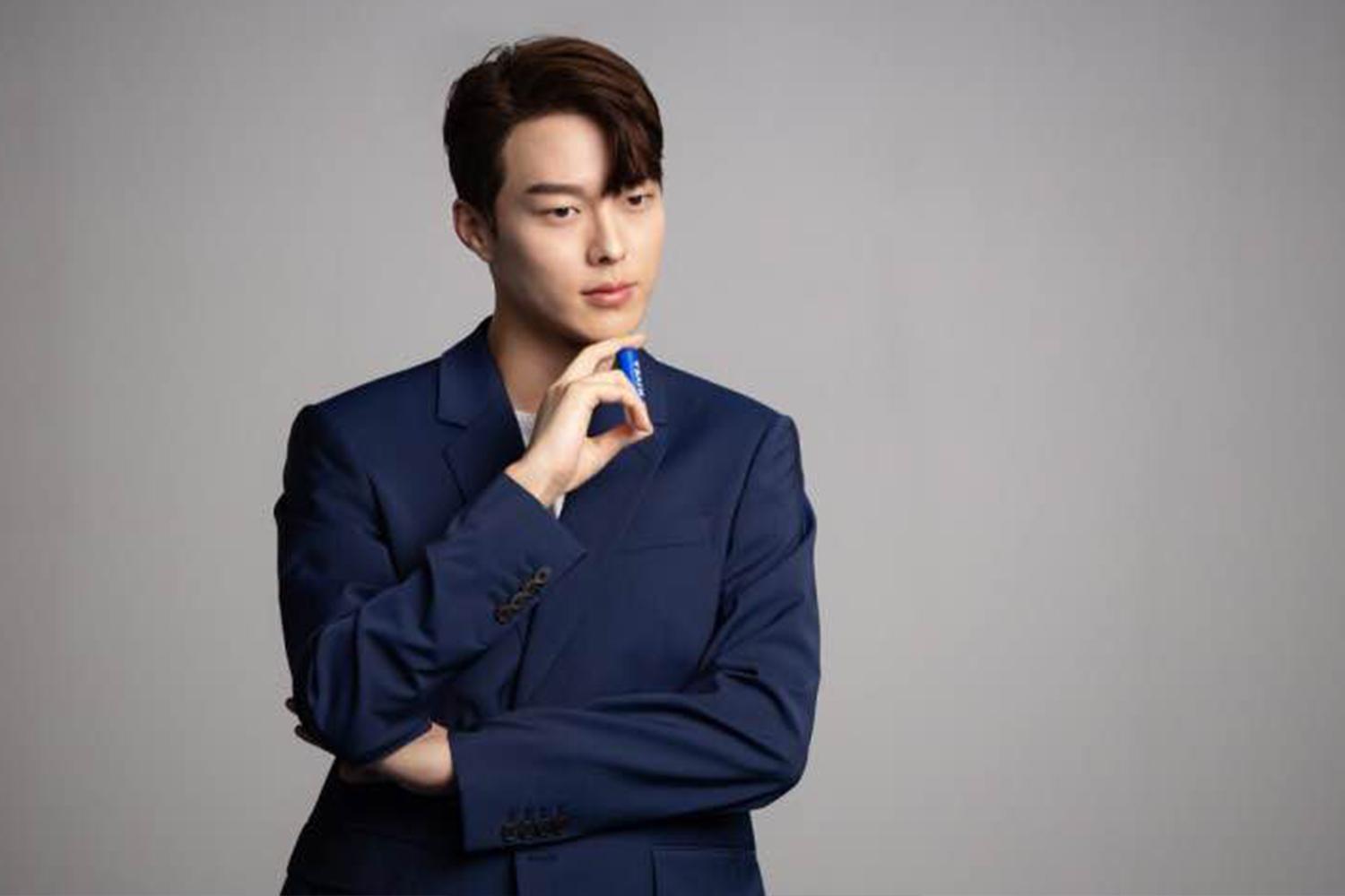 韓國男明星張基龍穿著藍色西裝，手託著下巴，背景簡約。