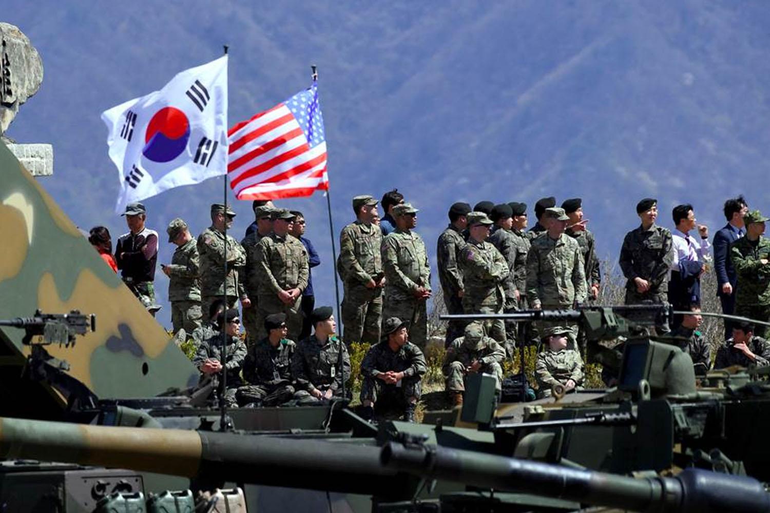Militari sudcoreani e americani in uniformi mimetiche in piedi di fronte a carri armati, con le bandiere della Corea del Sud e degli Stati Uniti sventolanti sullo sfondo.