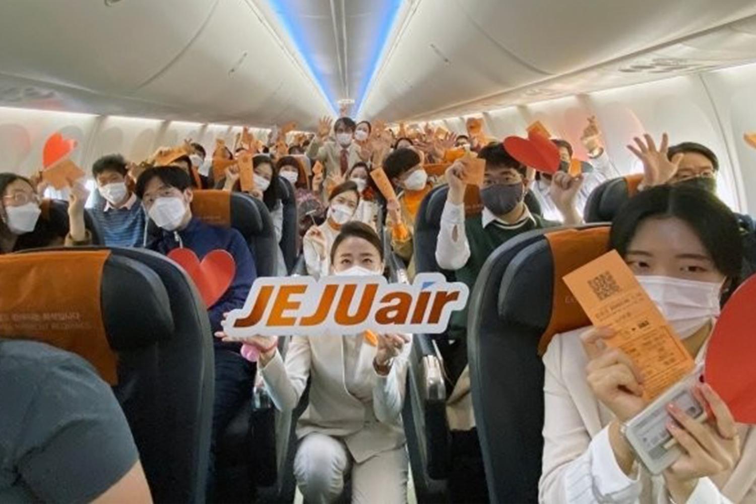成羣濟州航空乘客在機艙內，手持心形紙牌與JEJUair字樣，體驗飛行假期的樂趣。