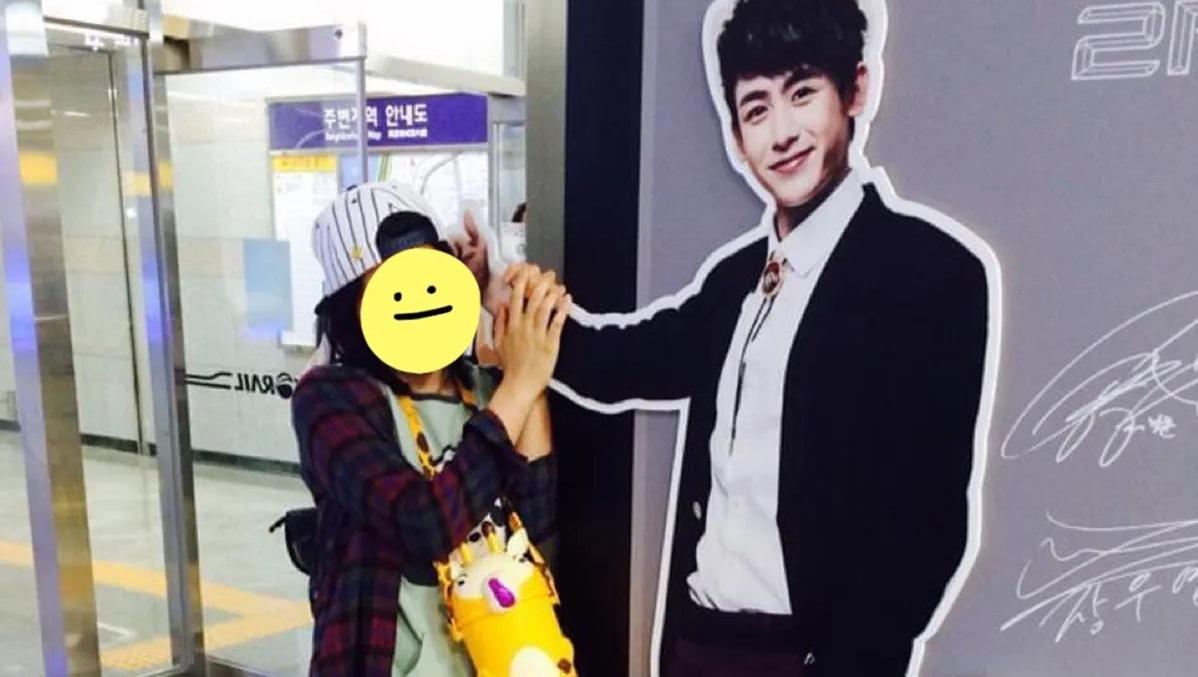 นักท่องเที่ยวถือของเตยจริงต่อหน้ารูปภาพขนาดใหญ่ของศิลปินเกาหลีในสถานีรถไฟฟ้าใต้ดิน