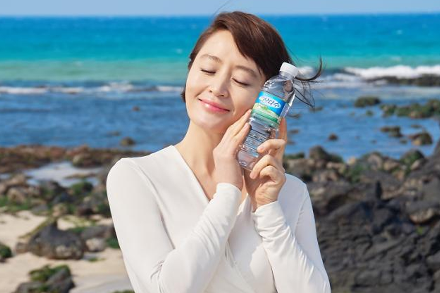 海灘背景下的礦泉水廣告，瓶子上印有韓文品牌名字，模特身穿白色上衣。