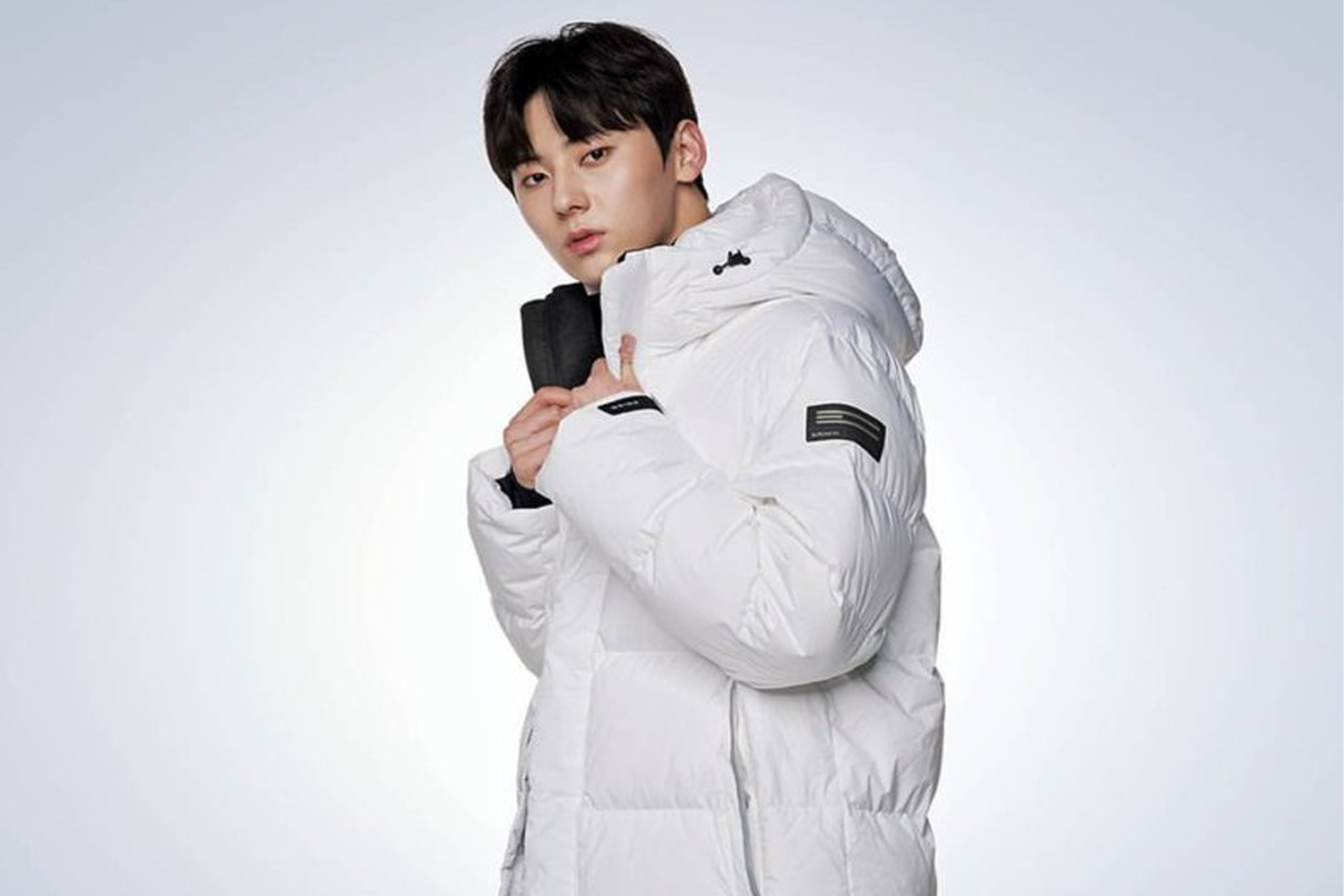 NU'EST成員旼炫穿著白色羽絨外套，站立在背景為灰色的牆前