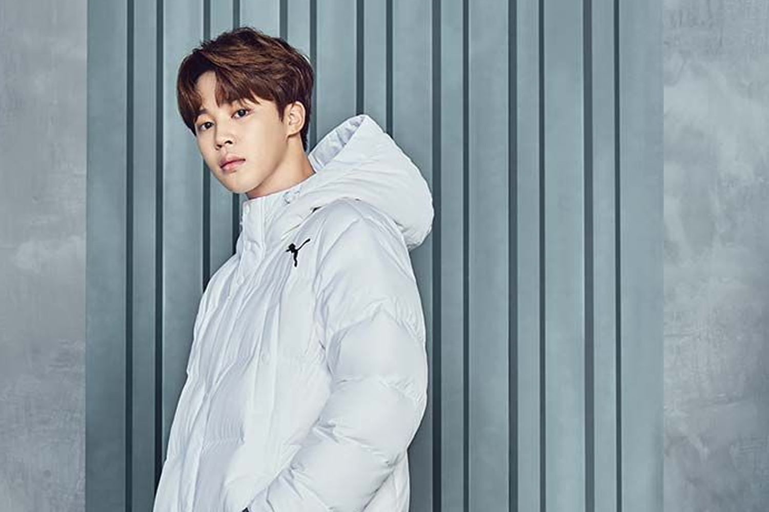 BTS成員Jimin穿著帶有品牌標誌的黑色羽絨外套，背景為藍天