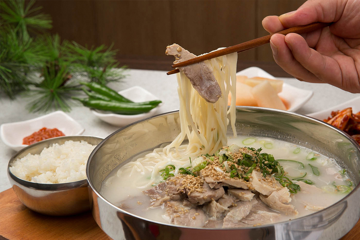 濟州著名料理豬肉湯麵的特寫，展示豐盛的湯麵和側邊的泡菜和米飯，象徵濟州的傳統美食及其多樣性。
