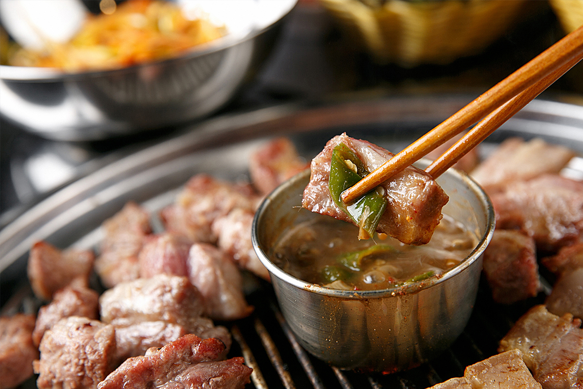 濟州黑豬肉烤肉的場景圖，有一片誘人的烤肉被筷子夾起，呈現濟州島獨具特色的美食魅力。