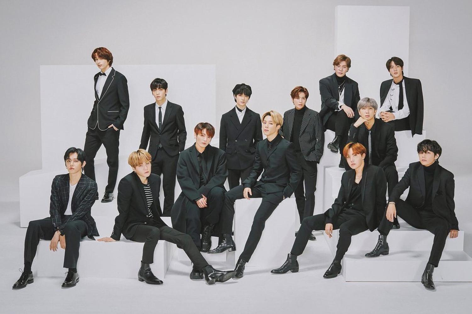 ภาพการรวมกลุ่มของศิลปินที่สวมชุดสูทสีดำในห้องสตูดิโอ, เซลบีเซอร์แบบเอาเท้ากางบางคนถือที่นั่งอยู่และบางคนยืนอยู่บนกล่องสีขาว พื้นหลังเป็นสีขาวสะอาดตา