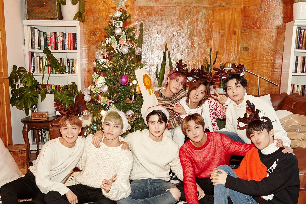 กลุ่มคนสวมเสื้อผ้าสบายๆ นั่งอยู่รอบต้นคริสต์มาสด้วยใบหน้าที่เบลอ ในห้องที่ตกแต่งด้วยต้นไม้และชั้นหนังสือเบื้องหลัง