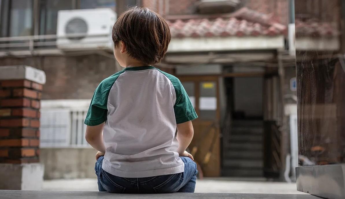 เด็กหนุ่มนั่งคุกเข่าอยู่ในบริเวณกลางแจ้ง บรรยากาศเงียบสงบ เห็นด้านหลังของเด็กหนุ่มกับพื้นหลังของตึกอิฐ