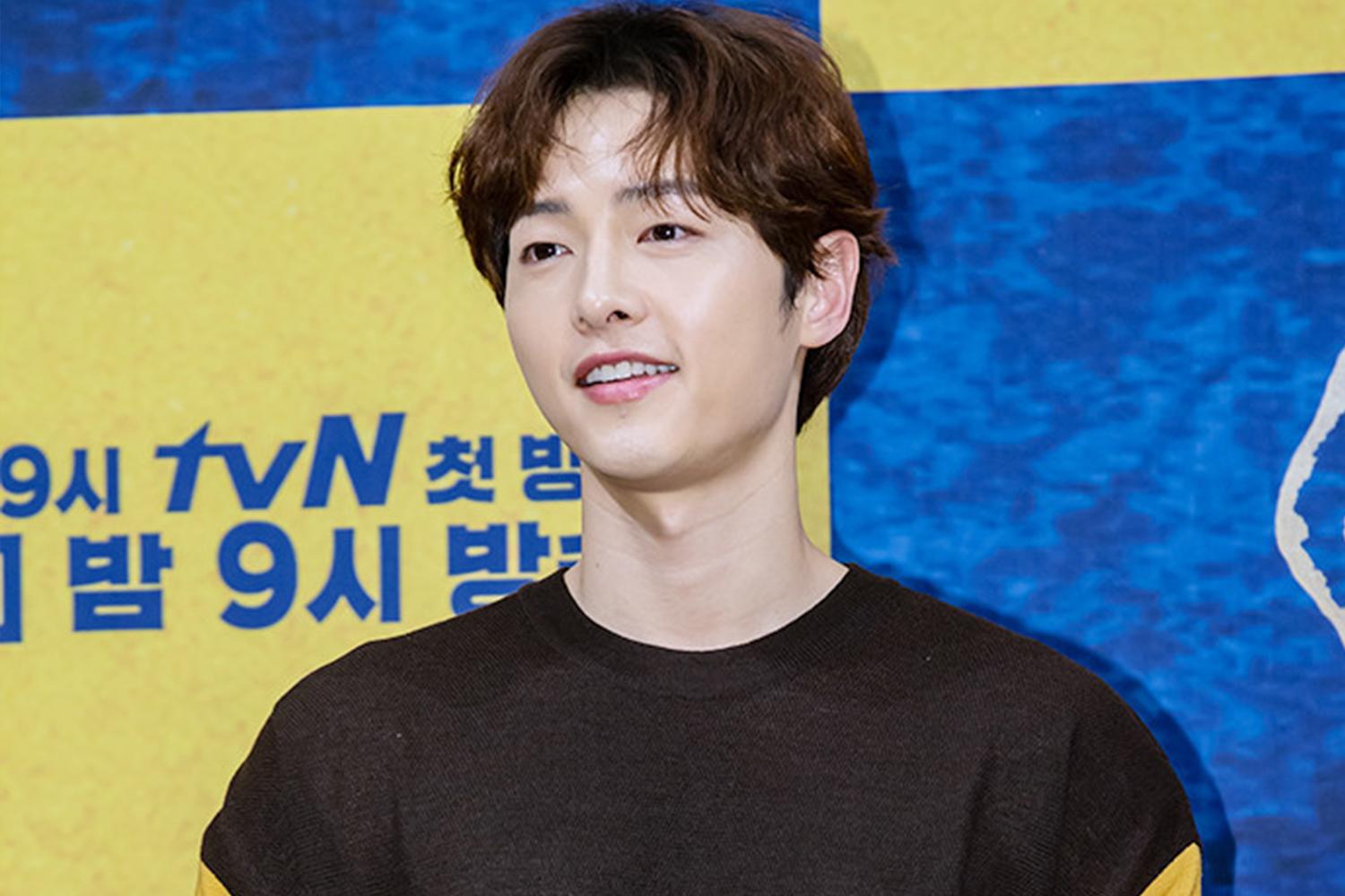 韓國著名男演員站在黃色背景前，他身穿黑色和黃色拼接毛衣，參與電視活動。