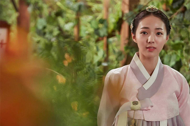 穿著傳統韓服的女性角色站在綠色背景前，背景有古色古香的裝飾，情景帶人進入《哲仁王后》的時代環境。