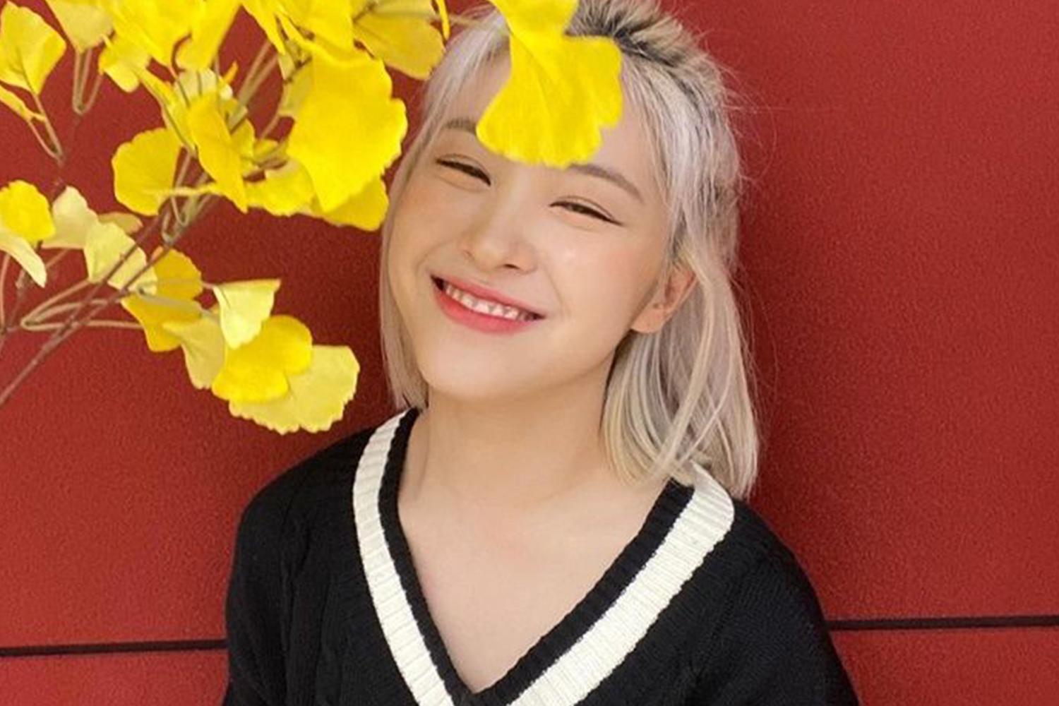 ITZY 成員留真站在紅色背景的牆邊，黃葉裝飾圖片右側，穿著黑塑蠶衣。