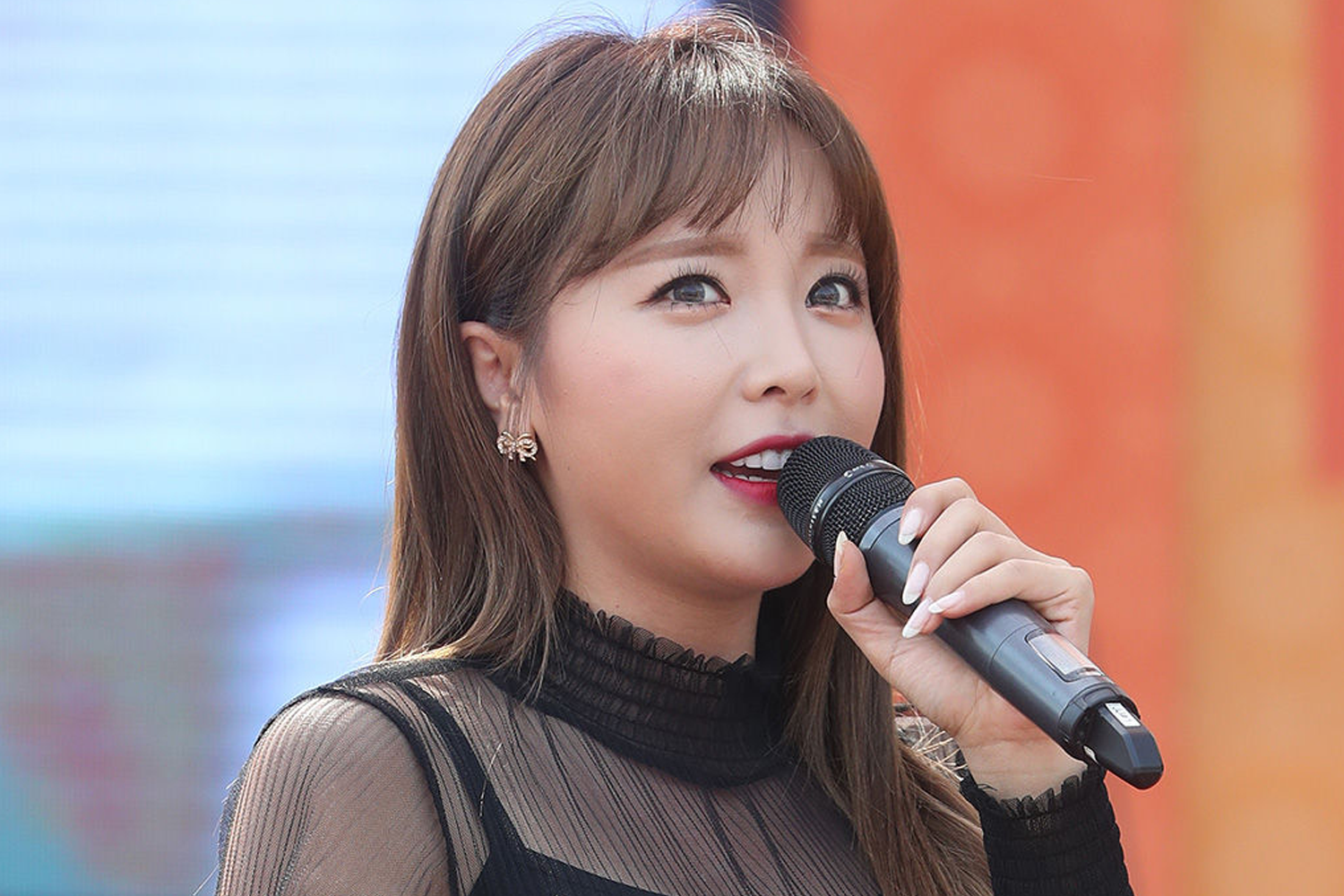 韩国知名Trot歌手洪真英手持麦克风，穿着黑色透明服装在户外舞台上演出，背景颜色鲜艳。