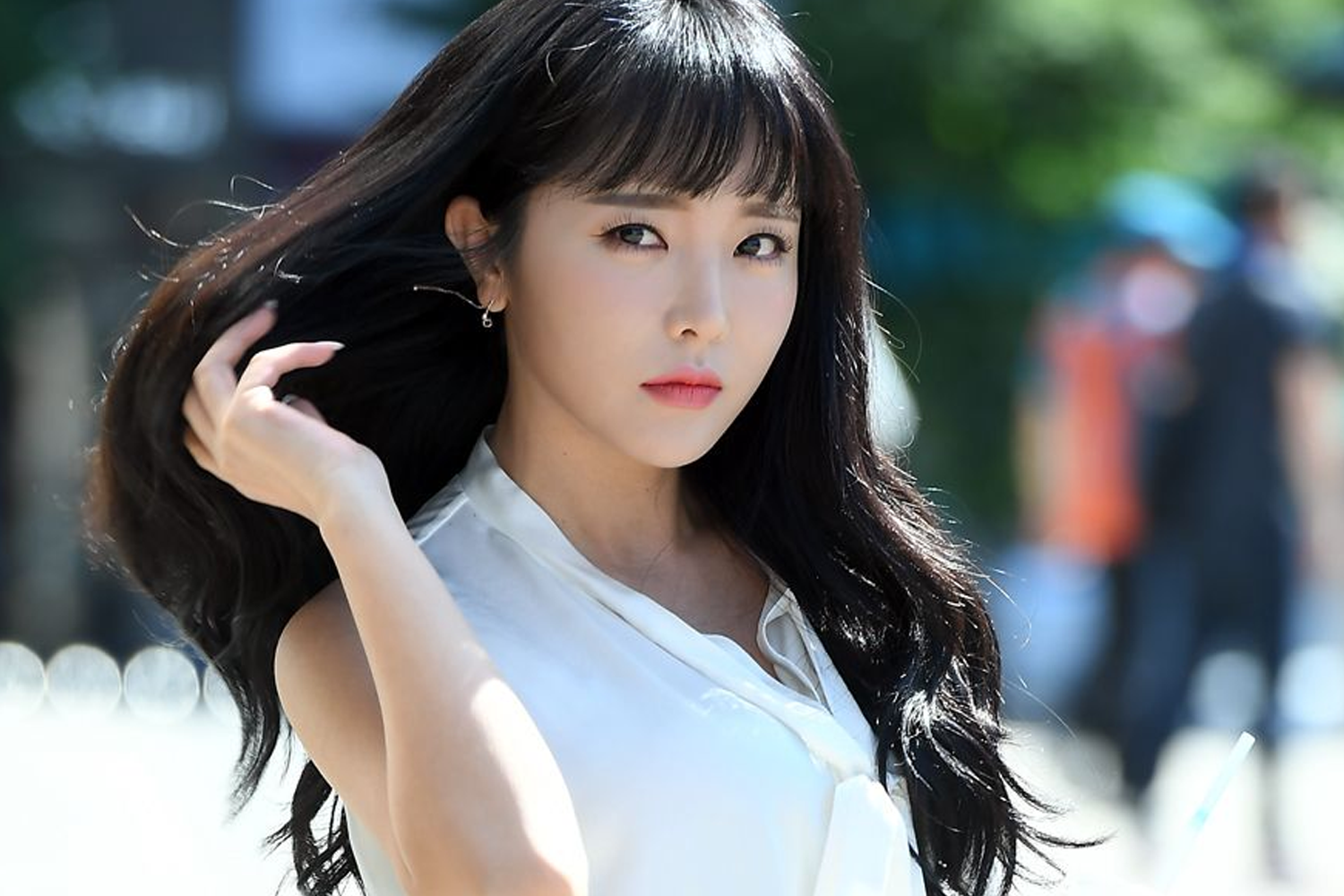 韩国知名Trot歌手洪真英身穿白色服装，手握黑色长发，背景模糊的人影与绿色植物场景。