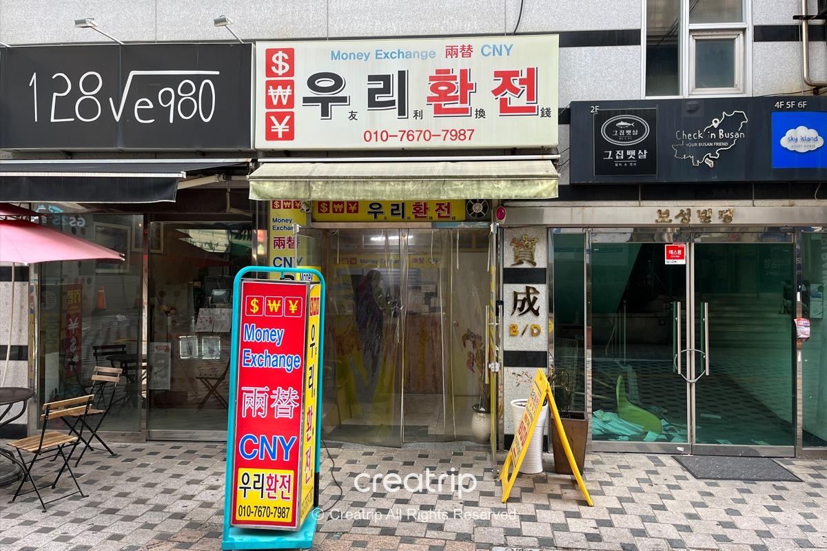 釜山南浦洞友利換錢所的入口，標示出可兌換的貨幣，店面前擺放著兌換相關的指示牌。