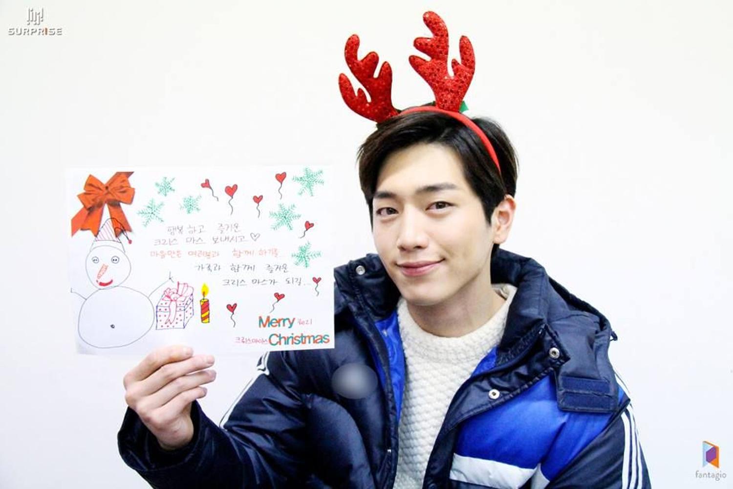 韓國藝人頭戴紅色麋鹿角看板，手持聖誕賀卡，背景簡約而富有節日氣氛。
