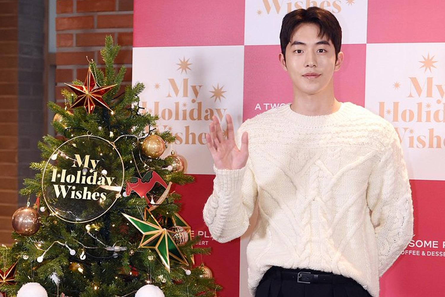韓國藝人身穿白毛衣，在聖誕樹旁揮手微笑，背景為My Holiday Wishes的活動看板。