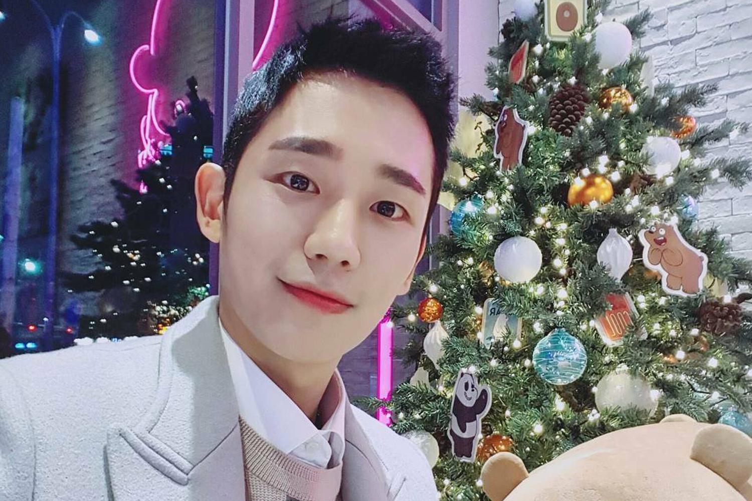 韓國藝人身穿淺色大衣，背景有裝飾華麗的聖誕樹，展現時尚感。