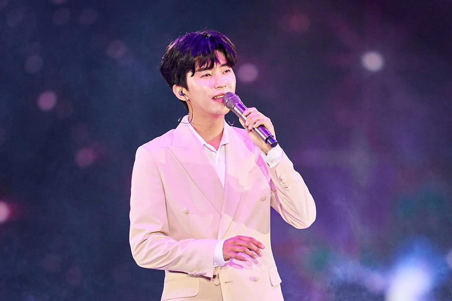 穿著粉色西裝的韓國男藝人在舞台上演唱，背景為夢幻的燈光效果。