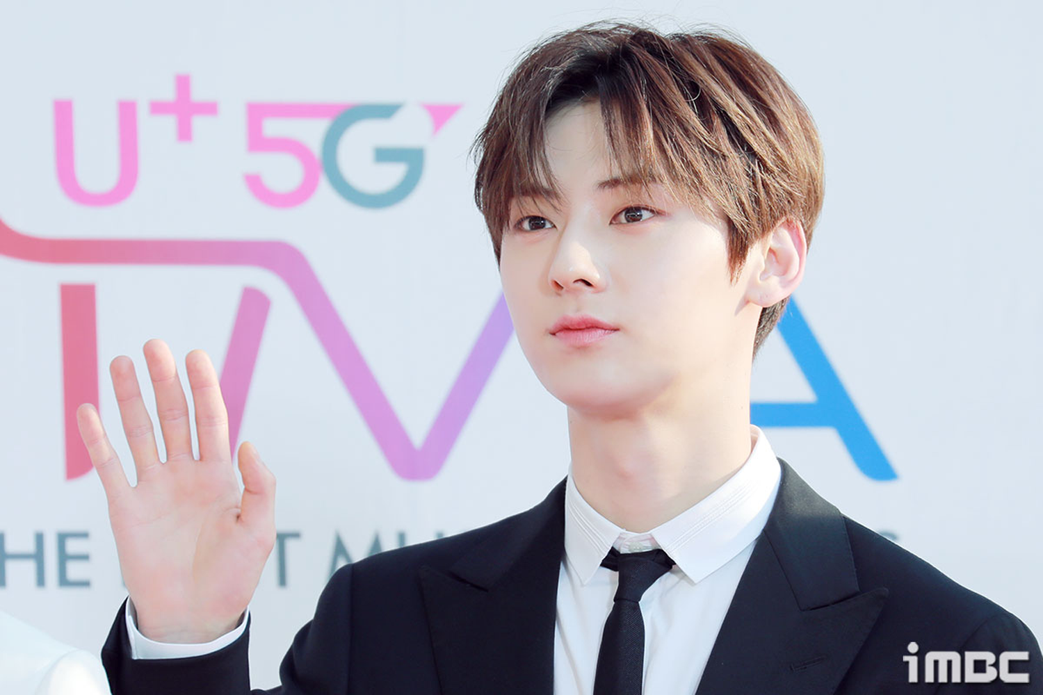 韓國男子組合NU'EST的成員旼炫，身穿黑色西裝和白襯衫，背景為紅色地毯活動現場