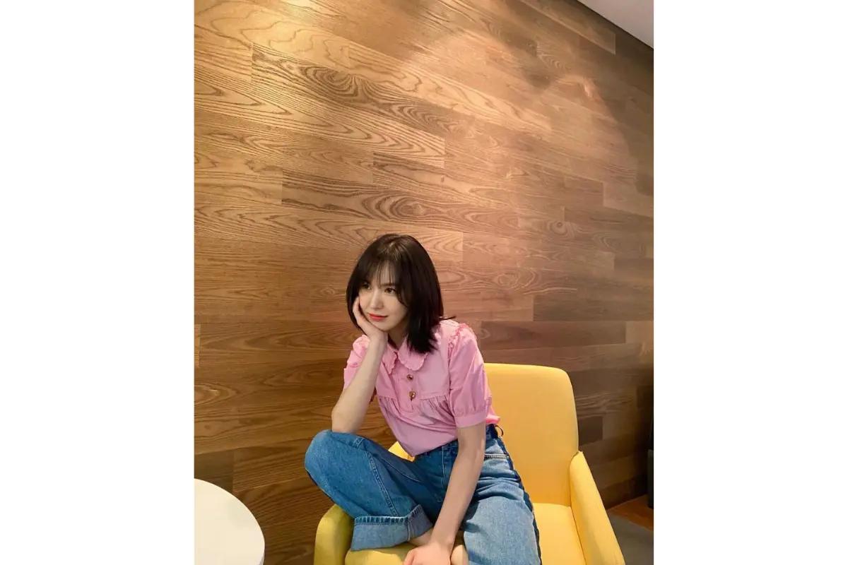 韓国のハッシュカットスタイルを着た女性が、木目調の壁を背景に、ピンクのシャツとデニムジーンズを着て黄色の椅子に座っている様子。