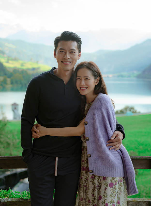 Actores Hyun Bin y Son Ye-jin detrás de cámaras del drama 'Crash Landing On You' con un fondo escénico junto al lago.