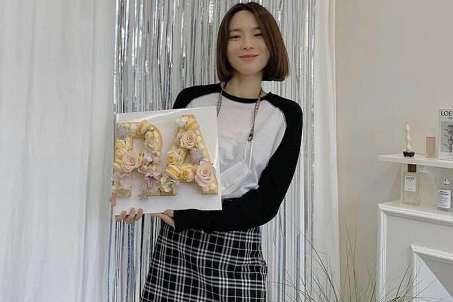 一位女士手持設計藝術字母嘅花牌,背景係一個裝飾有光面布幔嘅房間,環境時尚與精緻。