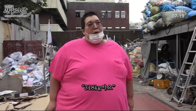 故Big Joeが屋外に立っている様子。彼はピンクのシャツを着て、現在の体重が318kgであることを示す字幕がスクリーンに表示されている。彼の健康問題にも焦点が当てられたシーン。