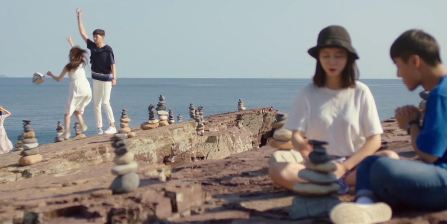 ชายหนุ่มและหญิงสาวกำลังเพลิดเพลินกับวิวทะเลที่บริเวณชายหาดของเกาหลีใต้ ที่มีการวางกองหินซ้อนกันอย่างสวยงาม