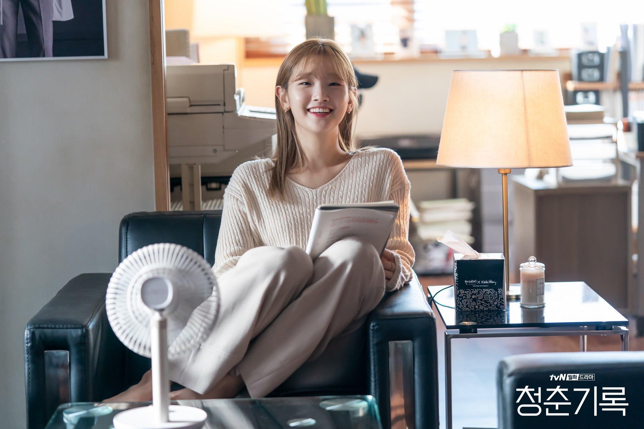 ผู้หญิงนั่งอยู่บนโซฟาในออฟฟิศขณะอ่านหนังสือ มีโคมไฟและพัดลมตั้งอยู่ข้าง ๆ ให้บรรยากาศการทำงานที่ผ่อนคลาย