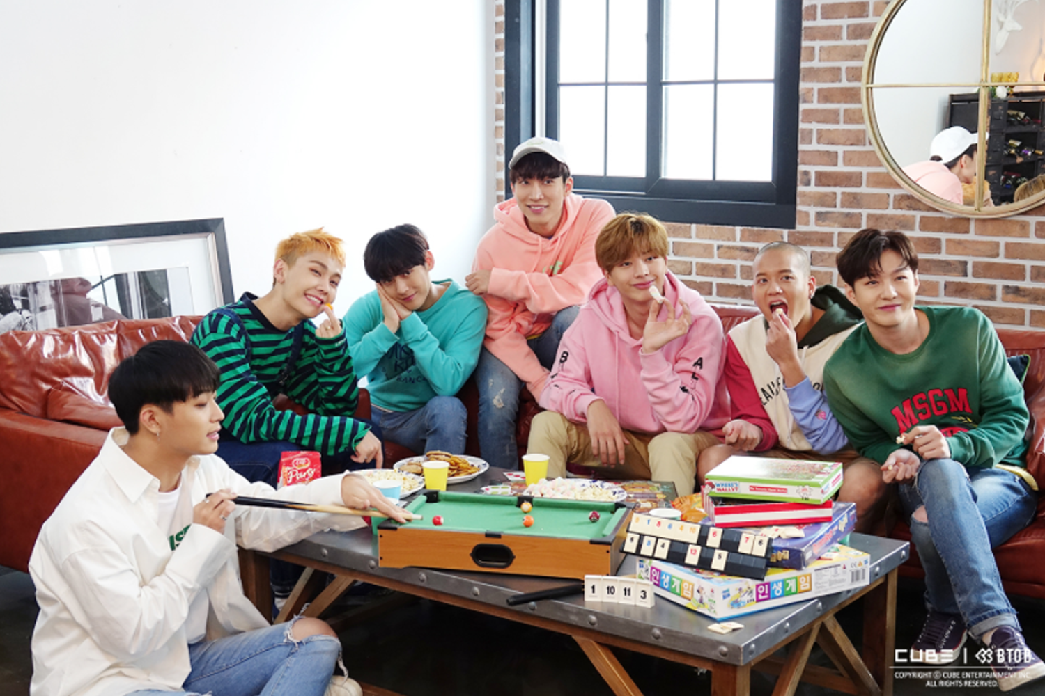 七位男團成員放鬆休息，圍坐在圓桌上，桌上擺放有飲料和食物，畫面呈現家庭與友好的社交場景。