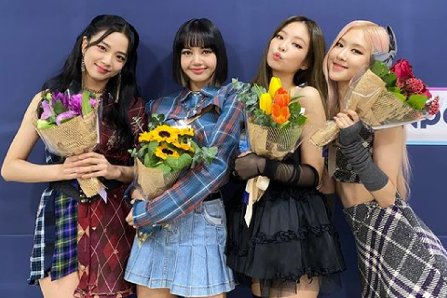 四位女子偶像成員手持花束站在一起，背景為淺藍色牆面，她們穿著色彩豐富的服飾，展現青春活力。