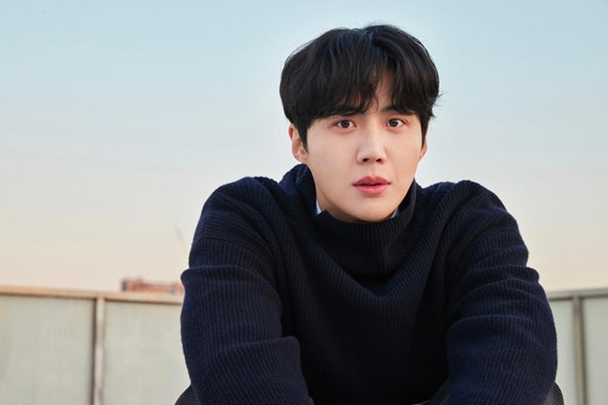 韓國演員金宣虎穿著淺色毛衣,坐在椅子上,背景是淺色的牆面,手託著下巴。