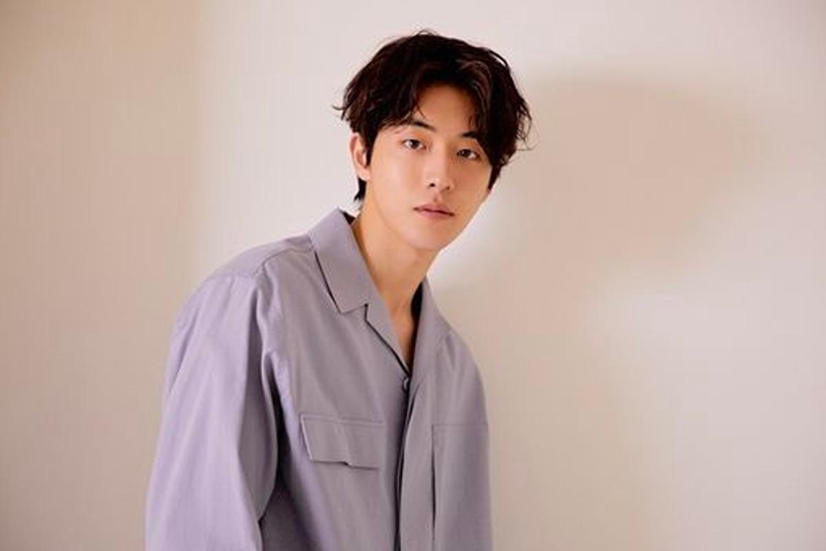 韓國演員南柱赫穿著白色T恤，坐在光亮的室內空間，背景為模糊的綠色背景牆。