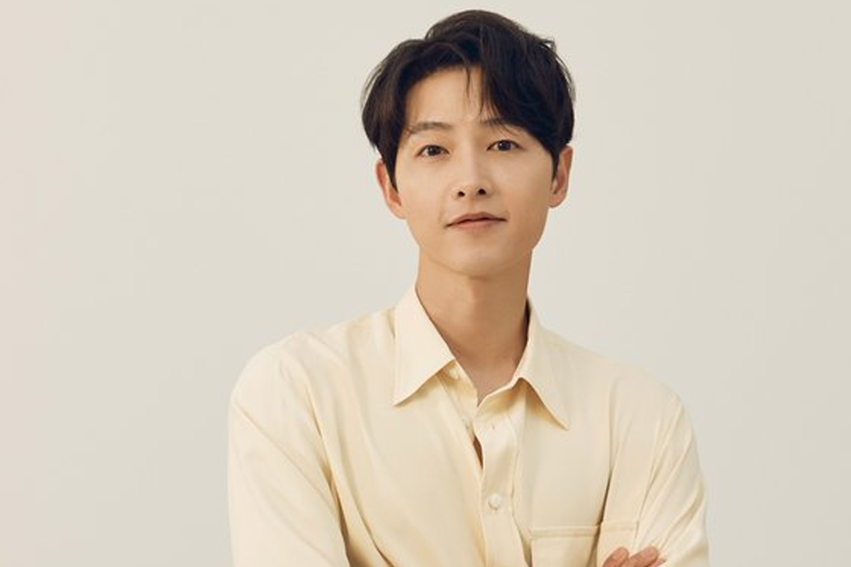 韓國演員宋仲基出席活動，穿著白色襯衫，背景為印有標誌的黑色牆壁。